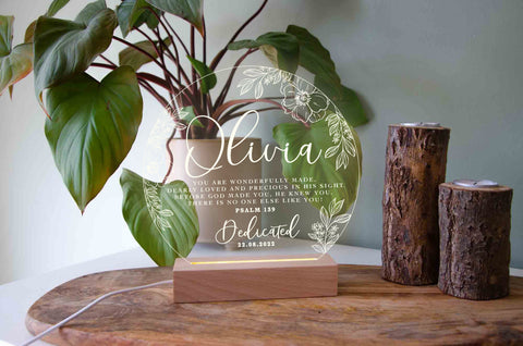 A personalised, transparent acrylic plaque with white cursive and print lettering stands on a wooden base. The personalised name is prominently featured in the centre in large cursive script. Surrounding the name is a circular floral and leaf design, with the text  Psalm 139 written below it. Below the verse, there’s a space for your custom event name and date.